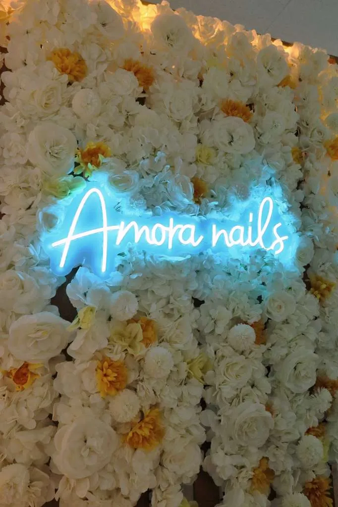 The Amora Nails neon sign glowing on a white floral wall inside the studio
